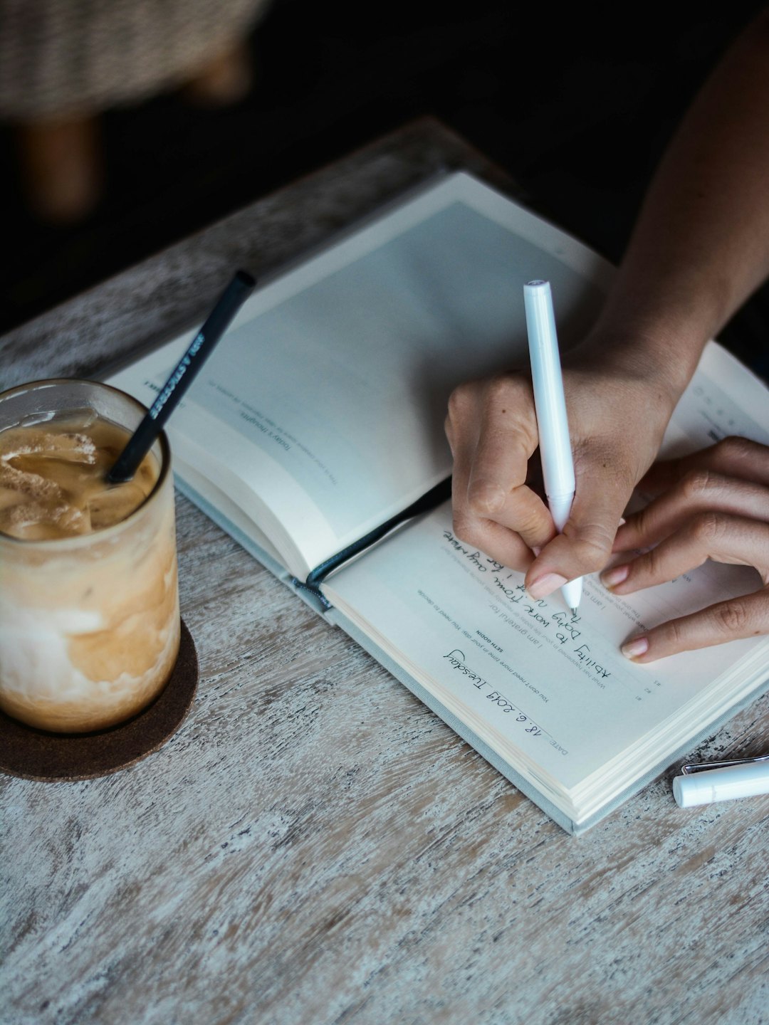 Manos escribiendo en un diario con un marcador blanco y café helado.