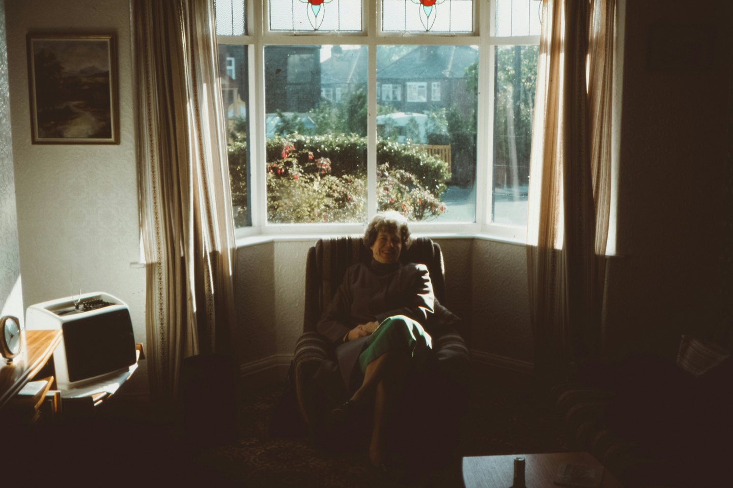 Una anciana sonríe sentada en un sillón junto a una ventana.